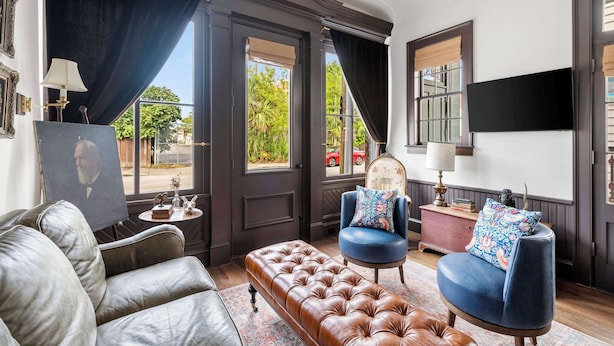 Una habitación con sofá de cuero, un sillón azul, un otoman marrón, un cuadro y una araña.