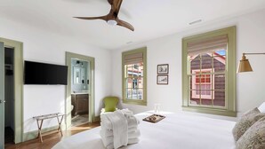 Habitación de hotel con una cama grande, ventilador de techo, televisión, una silla verde y ventana con vistas a edificios.