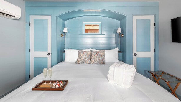 Un dormitorio con pared azul, cama blanca, dos almohadas, bandeja con vasos y plato, y televisor en la pared.