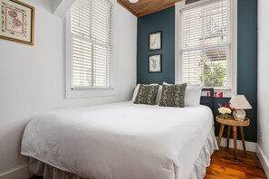 A bedroom with a large bed, two windows with blinds, a small bedside table with a lamp, and framed pictures on the wall. - Ebony & Ivory | Lofted Charm with Fireplace! (Charleston)