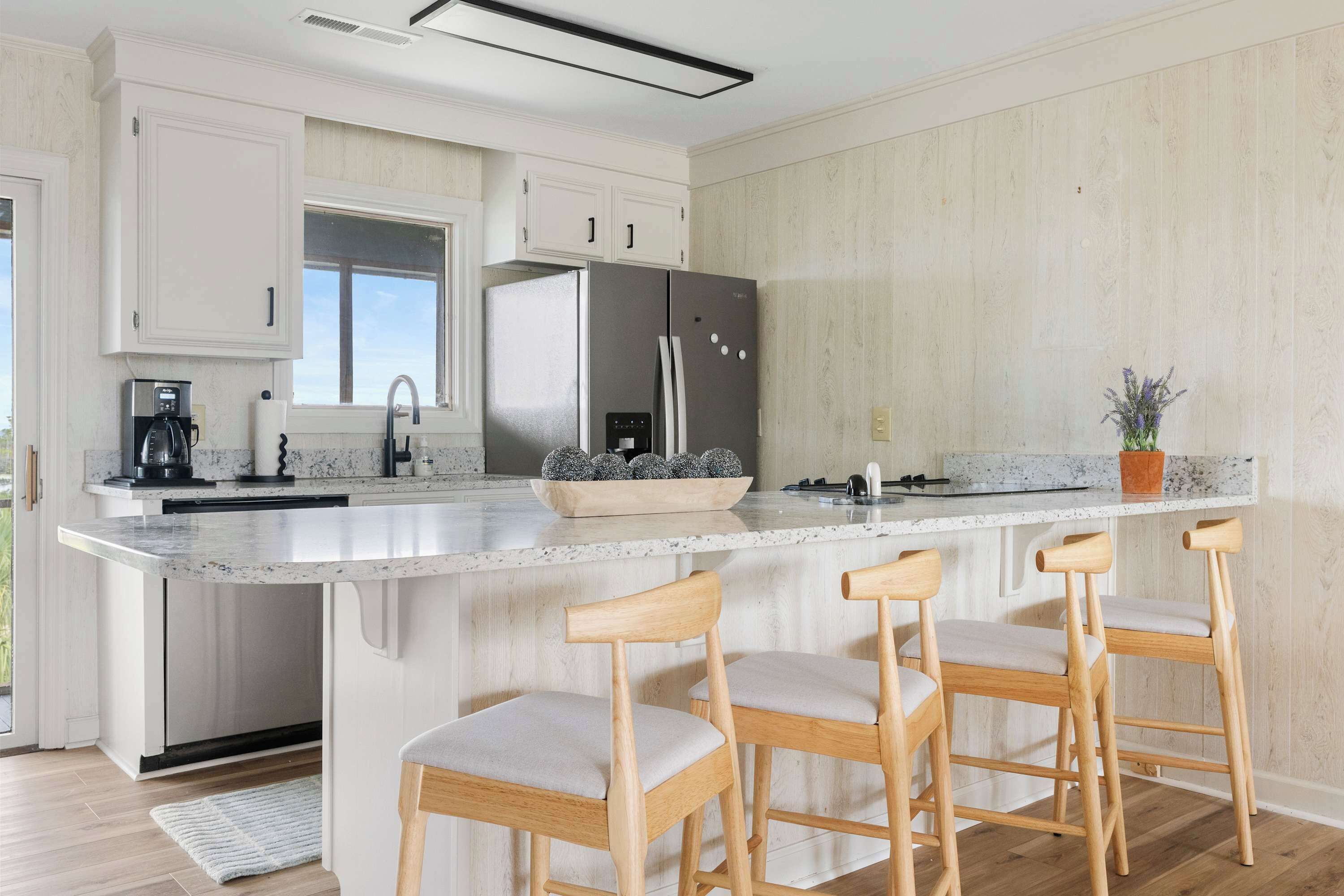 A modern kitchen with white cabinetry, a large island with bar stools, a stainless steel refrigerator, and a window providing natural light.