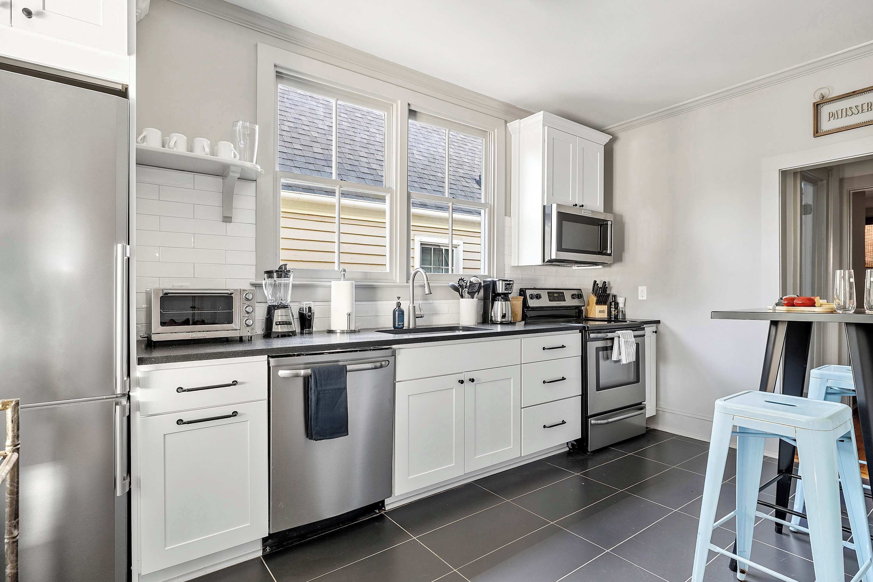 Una cocina moderna con armarios blancos, un refrigerador de acero inoxidable, un microondas y una estufa. Se ven dos ventanas que brindan luz natural, y un taburete pequeño al frente.
