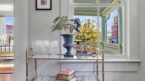 A room with a glass shelf holding wine glasses and books, a potted plant, and a view of a building through a window with blinds. - Incredible 4bd House, 1 block to King w. Parking! (Charleston)