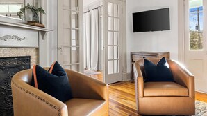 A living room with two leather armchairs, a fireplace, a mounted TV, and a large window. - Incredible 4bd House, 1 block to King w. Parking! (Charleston)