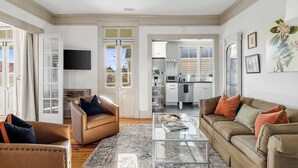 A living room with a sofa, armchairs, and a coffee table. There is a kitchen in the background with cabinets and a TV mounted on the wall. - Incredible 4bd House, 1 block to King w. Parking! (Charleston)