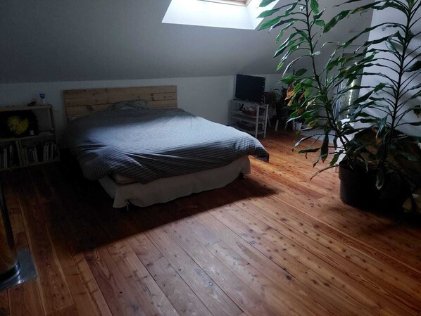 A bedroom with a wooden floor, a bed with a striped blanket, a wooden headboard, a small desk with a chair, a potted plant, and a skylight.