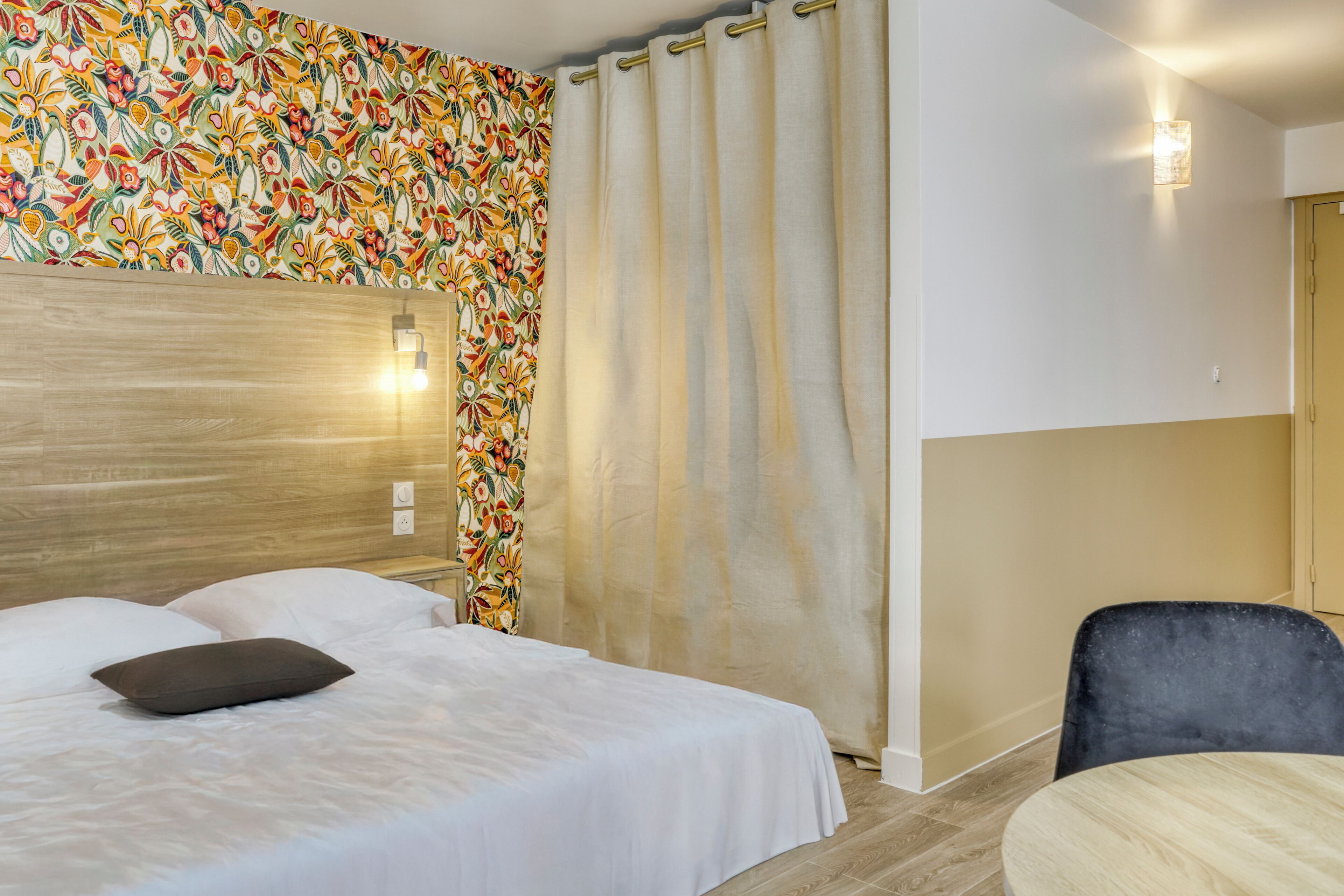 Una habitación de hotel con cama, una silla, una cortina y una pared con estampado floral.