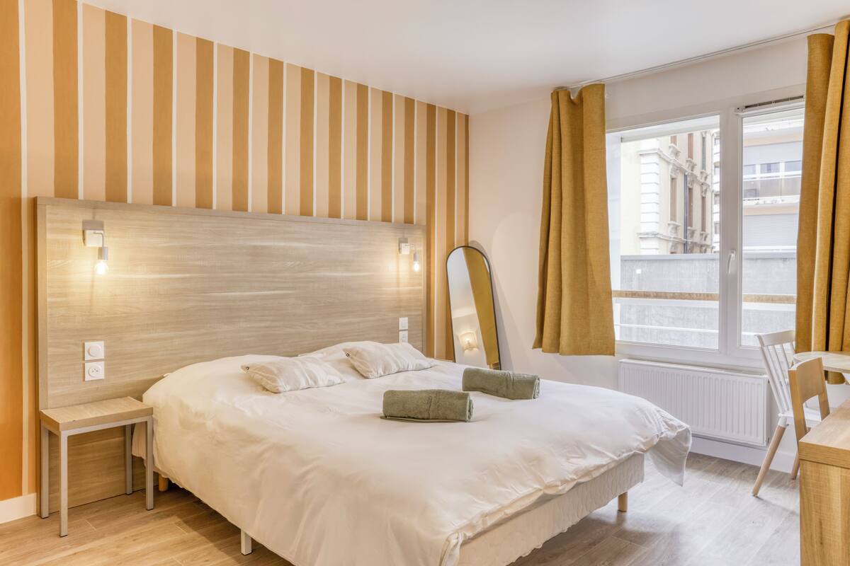 A hotel room with a large bed, a bedside table, a chair, a window with curtains, and a view of buildings outside.