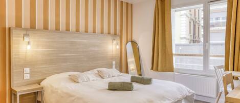 Une chambre d’hôtel avec un grand lit, une table de chevet, une chaise, une fenêtre avec des rideaux et une vue sur les bâtiments environnants.