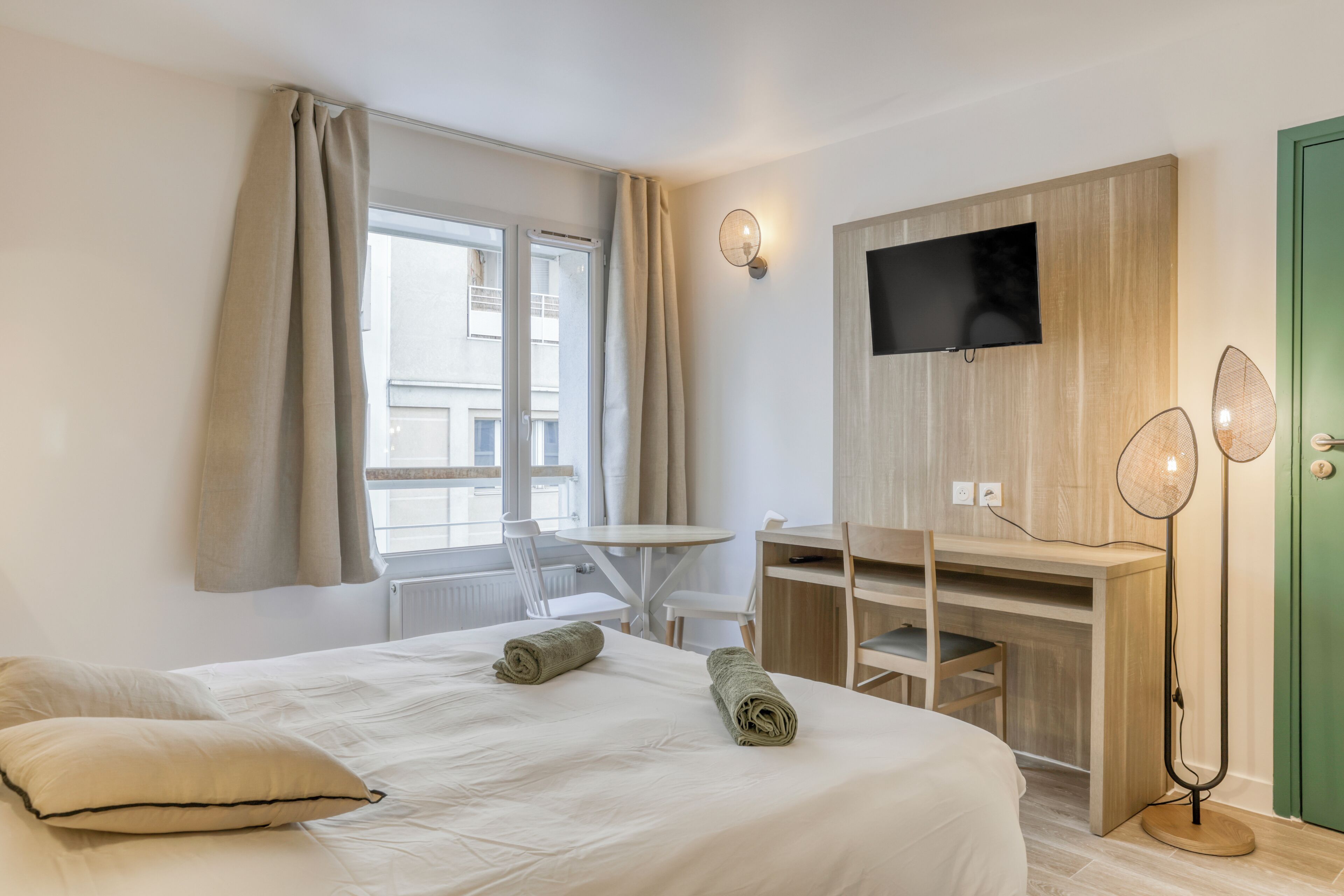 A hotel room with a bed, a desk, a chair, a TV, and a floor lamp.