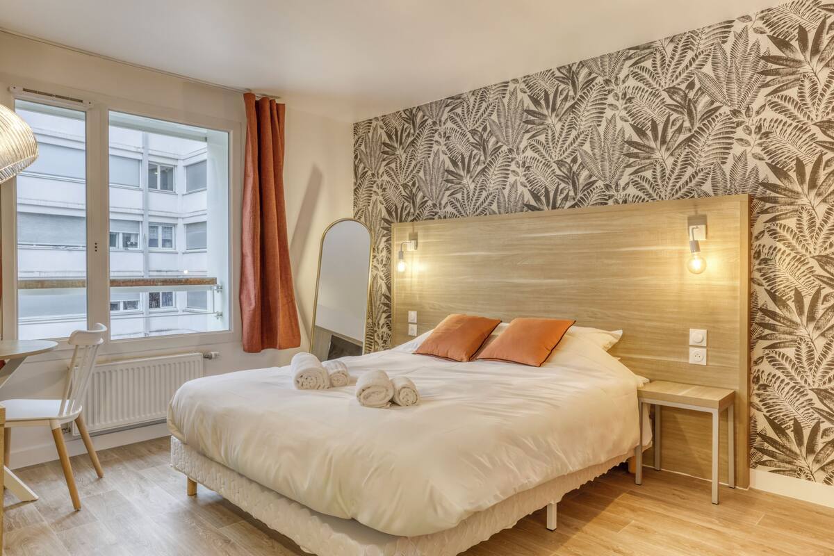 A hotel room with a large bed, a bedside table, a chair, and a view of a building through a window.