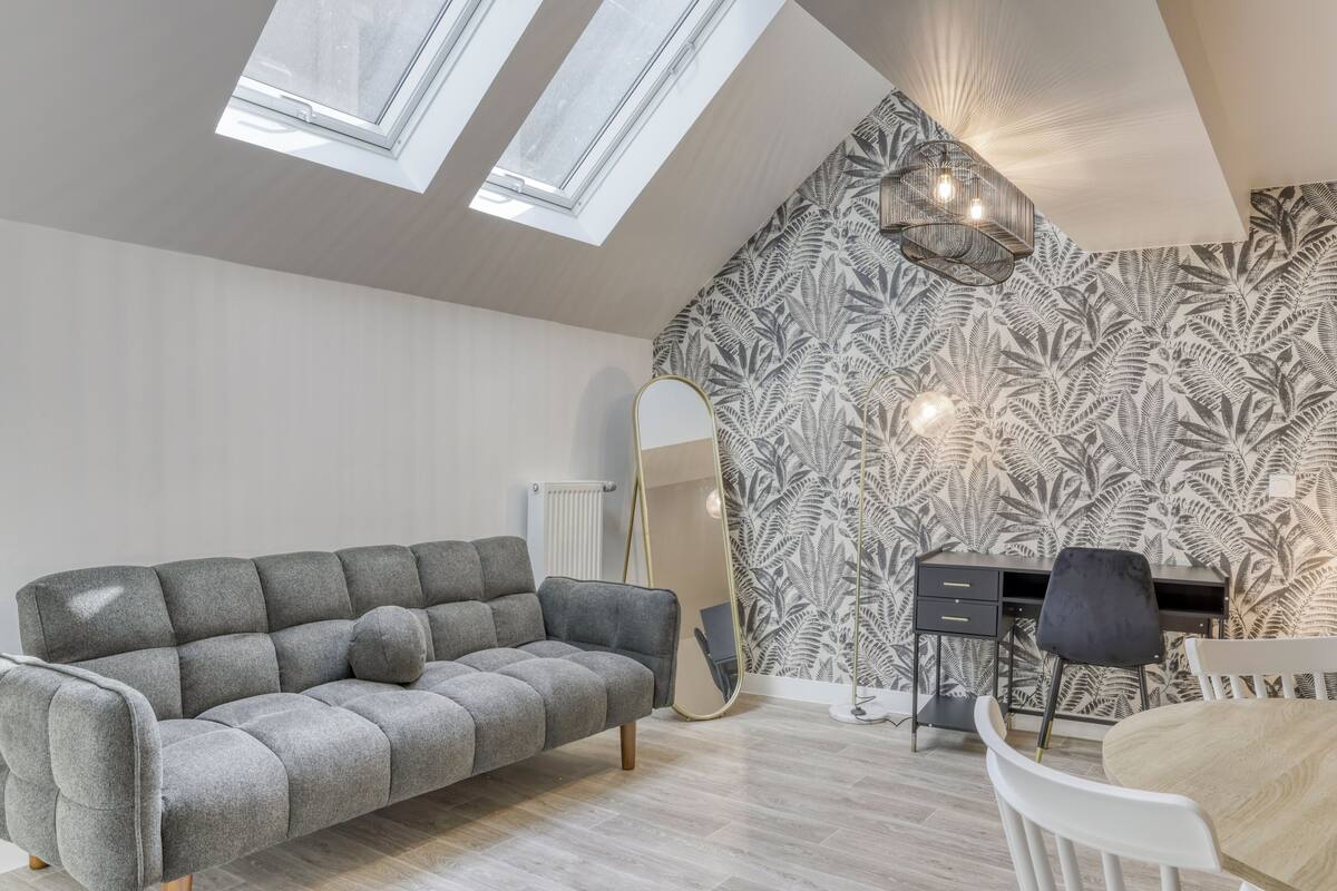 A modern living room with a grey sofa, a dining table, and a black desk. There are two skylights and a patterned wallpaper on the wall.