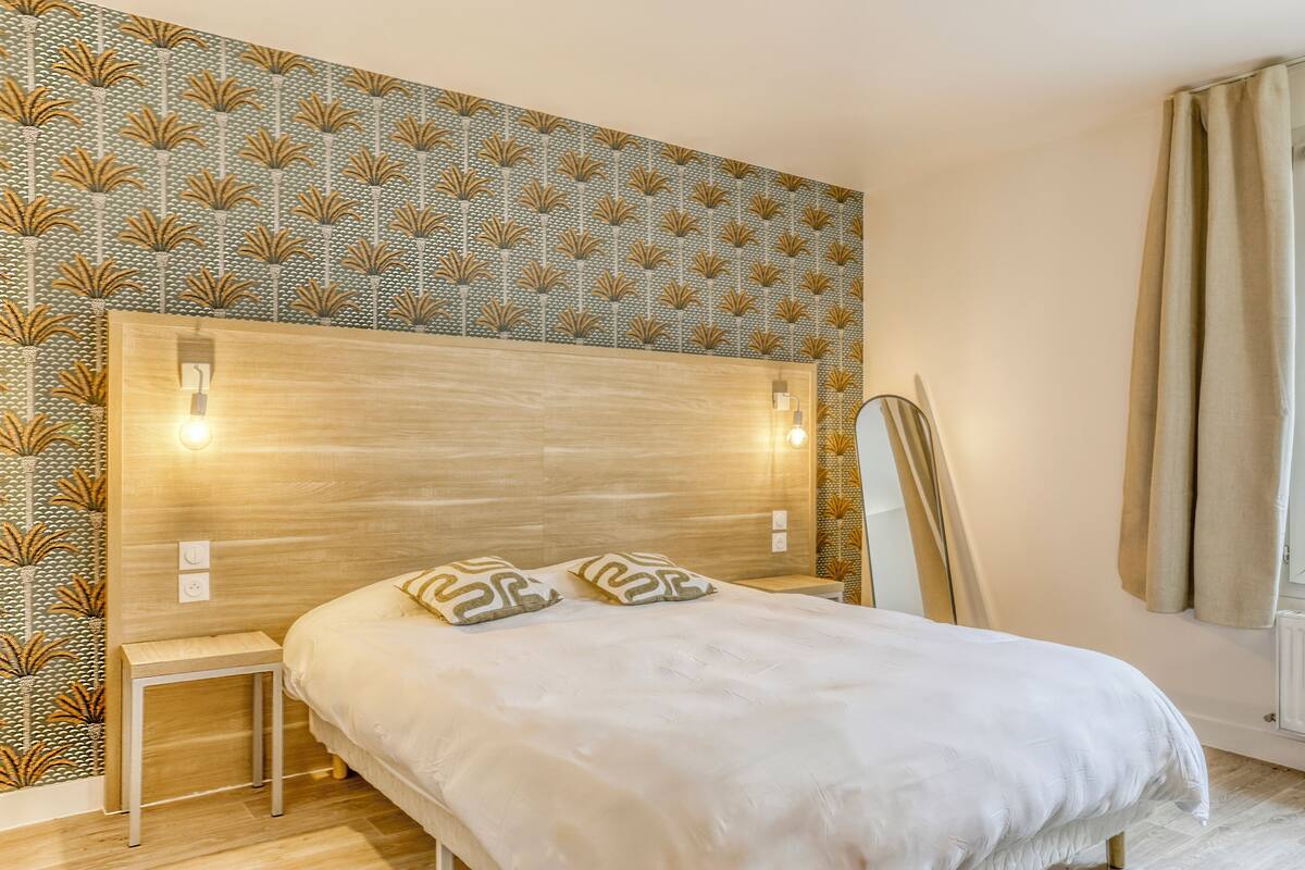 A bedroom with a patterned wallpaper, a wooden headboard, bedside lamps, and a bed with pillows.