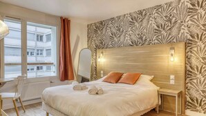 Hotelkamer met een groot bed, nachtkastje, stoel, raam met gordijnen en behang met een patroon.