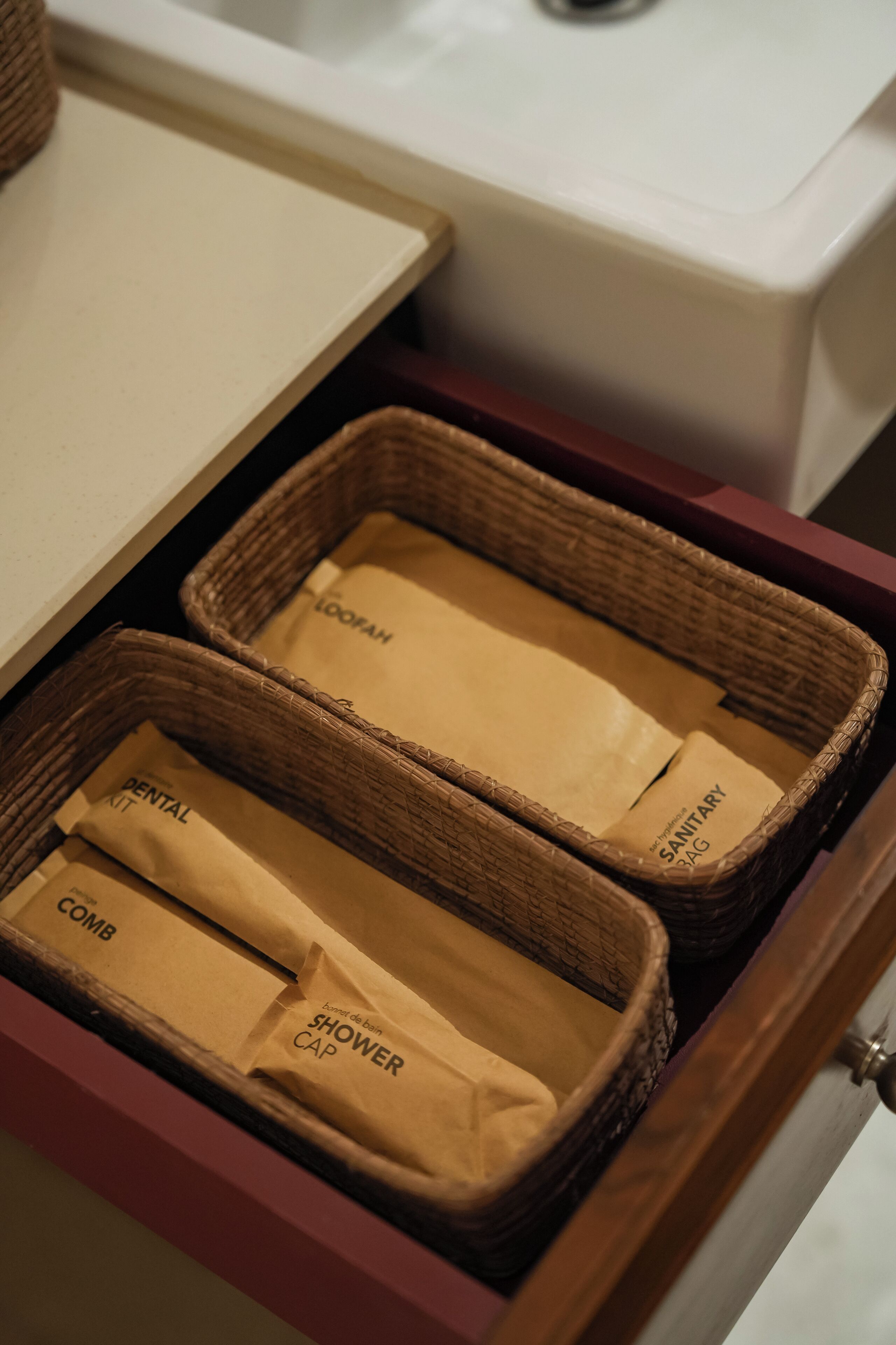 A drawer with wicker baskets containing labeled items such as a dental kit, comb, and shower cap.