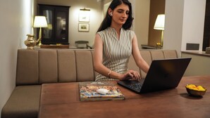 En person sitter vid ett träbord med en bärbar dator, en tidning och en skål med frukt.