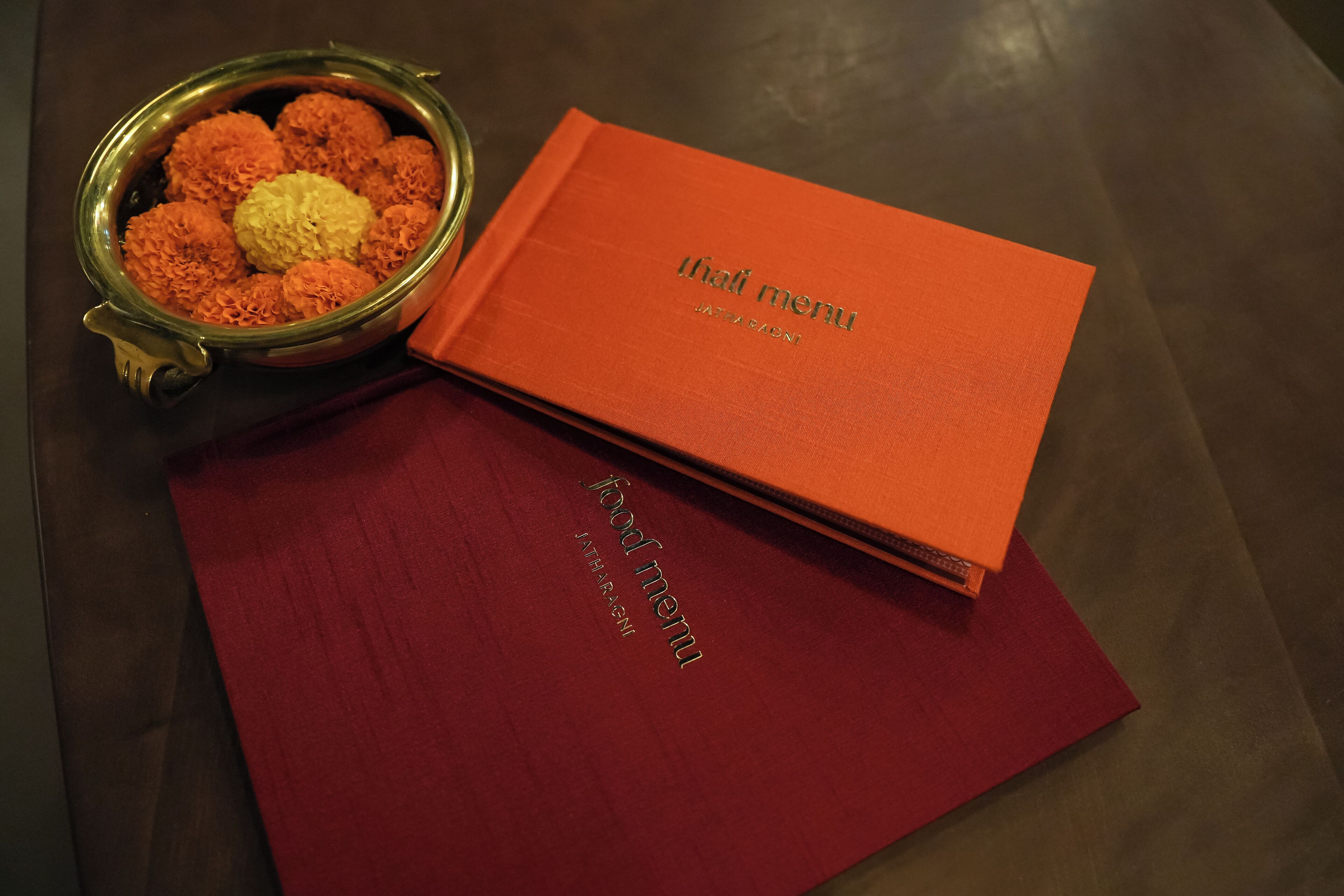 Een rood menuboek met de opdruk 'thai menu', geplaatst op een houten tafel naast een bloemenkom met goudsbloemen.