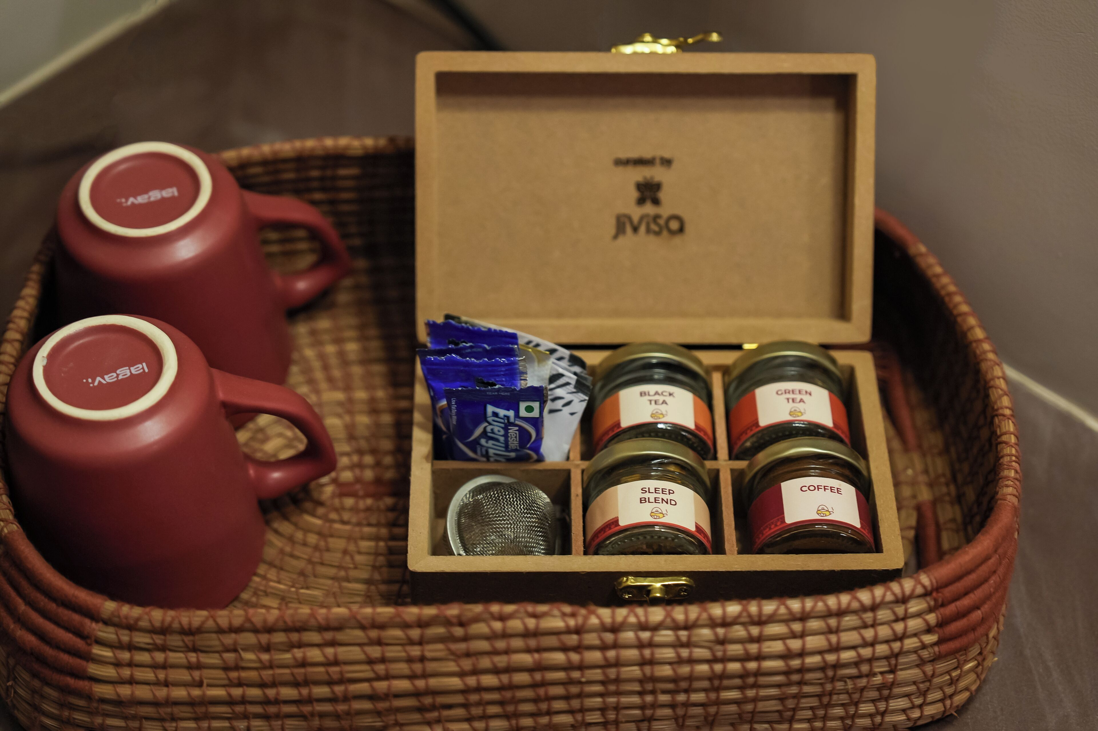 A wicker basket containing a tea set with two red cups, a tea bag, and three jars labeled 'SLEEP BLEND,' 'BLACK TEA,' and 'COFFEE.'