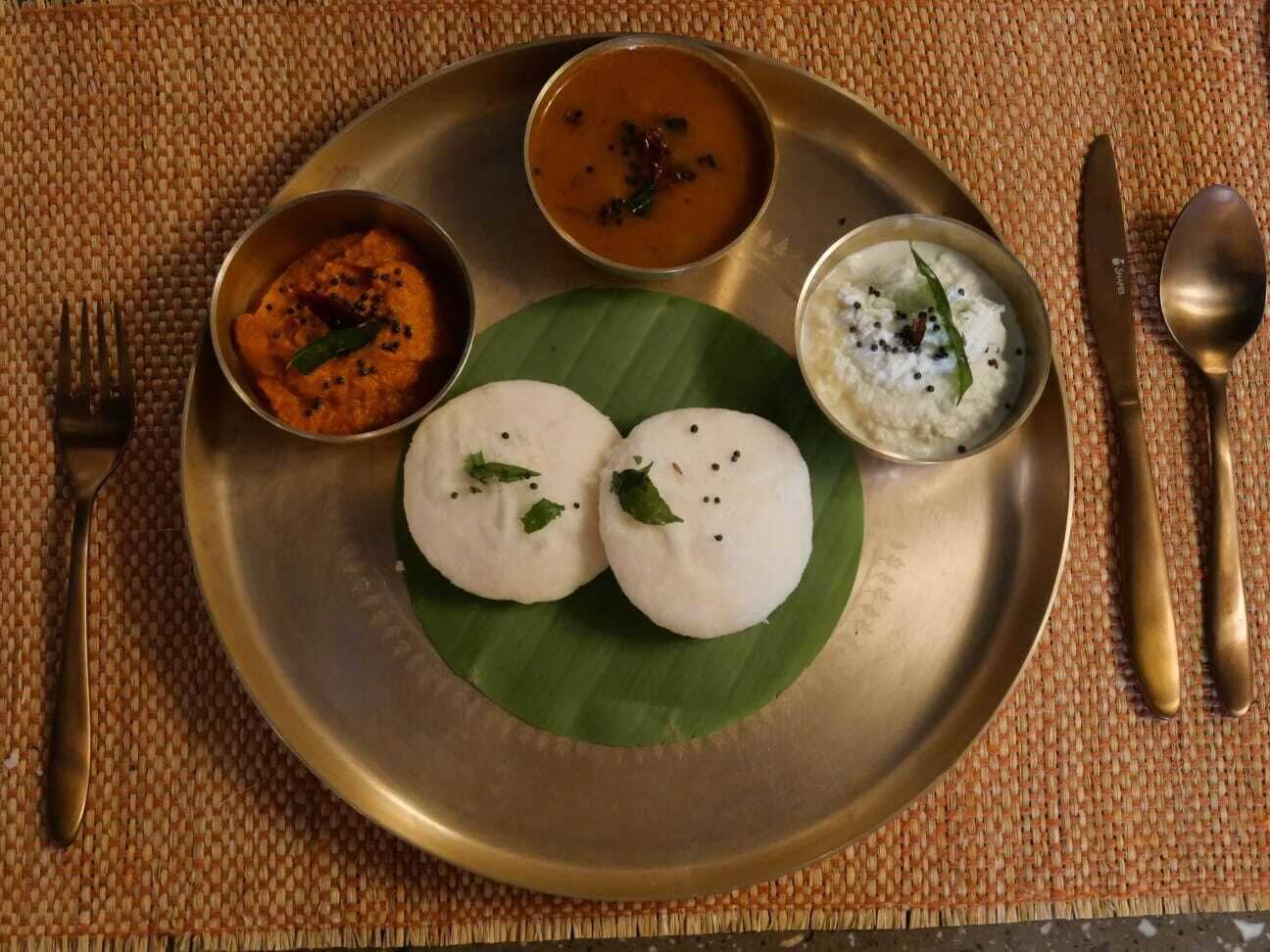 A traditional South Indian meal on a metal plate with two idlis, a bowl of sambar, and a bowl of coconut chutney.