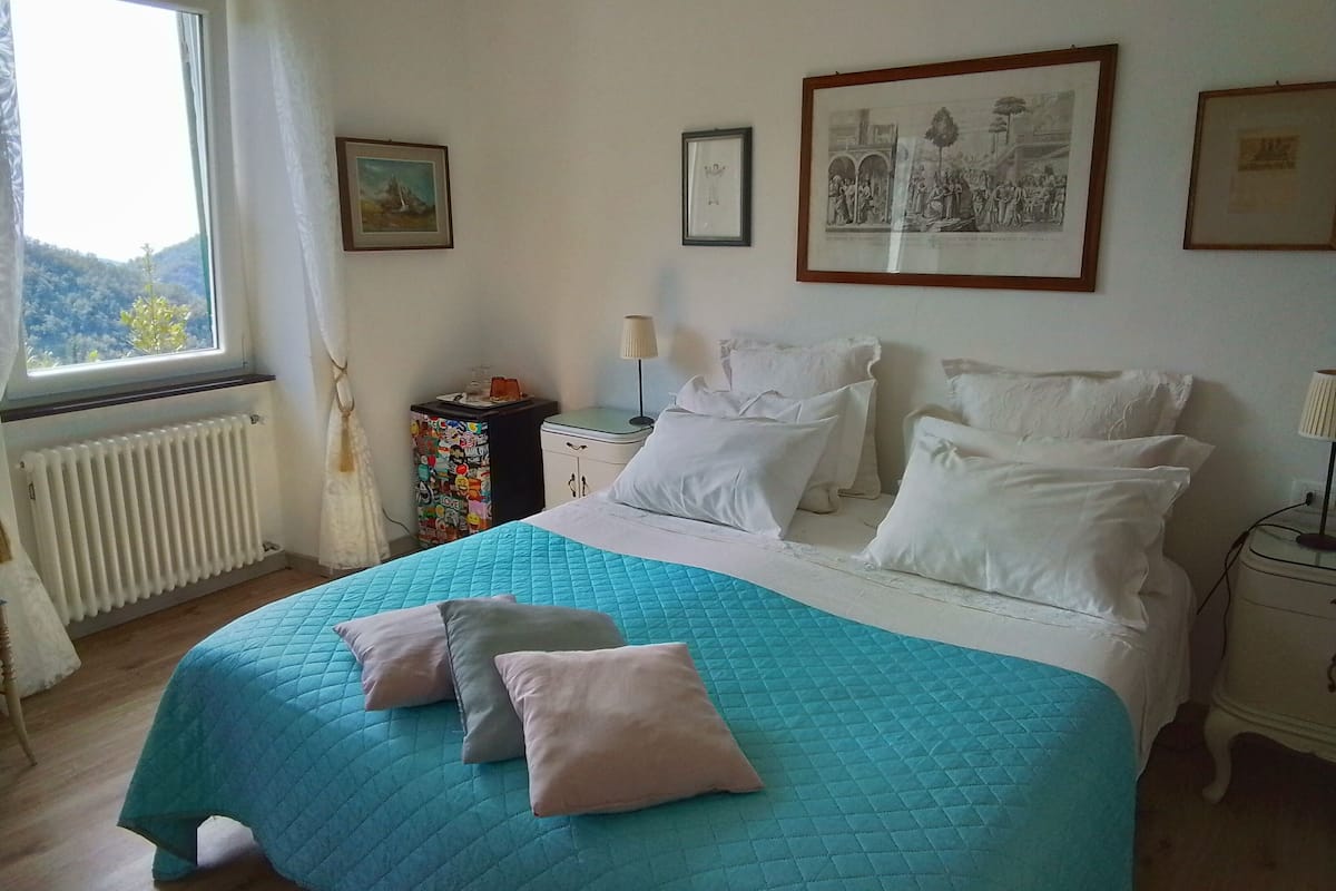 A bedroom with a bed, a window, a radiator, a small table, and framed pictures on the wall.