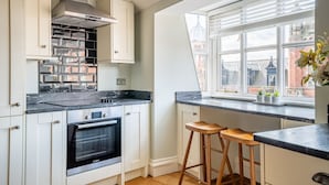 A modern kitchen with white cabinetry, a black countertop, and a built-in oven. There are two wooden stools and a window with a view of buildings. - Clifford St - Apartment 8 - sleeps 4 guests  in 2 bedrooms (York)