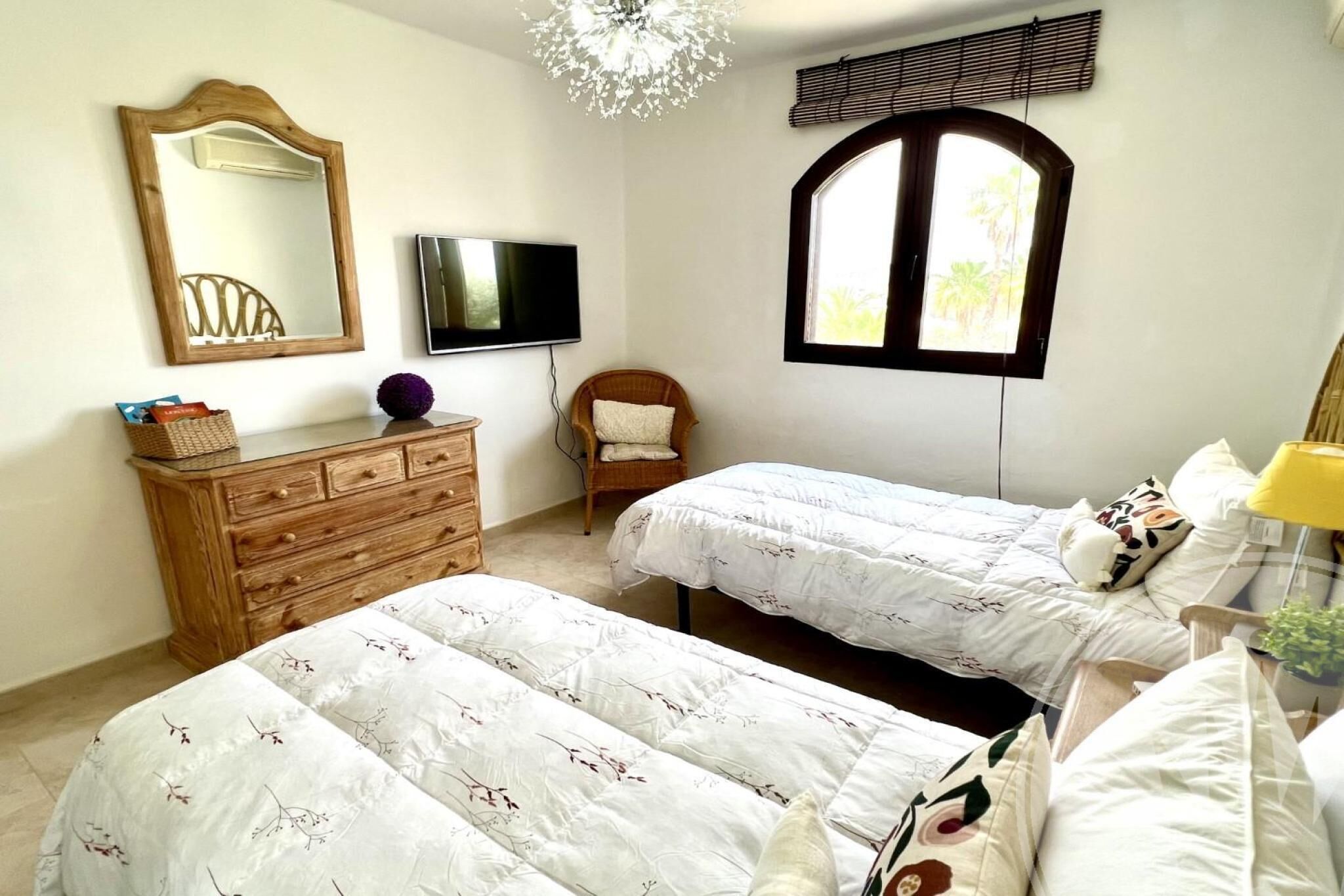 Een slaapkamer met twee bedden, een houten dressoir, een televisie, een raam met jaloezieën en een kroonluchter.