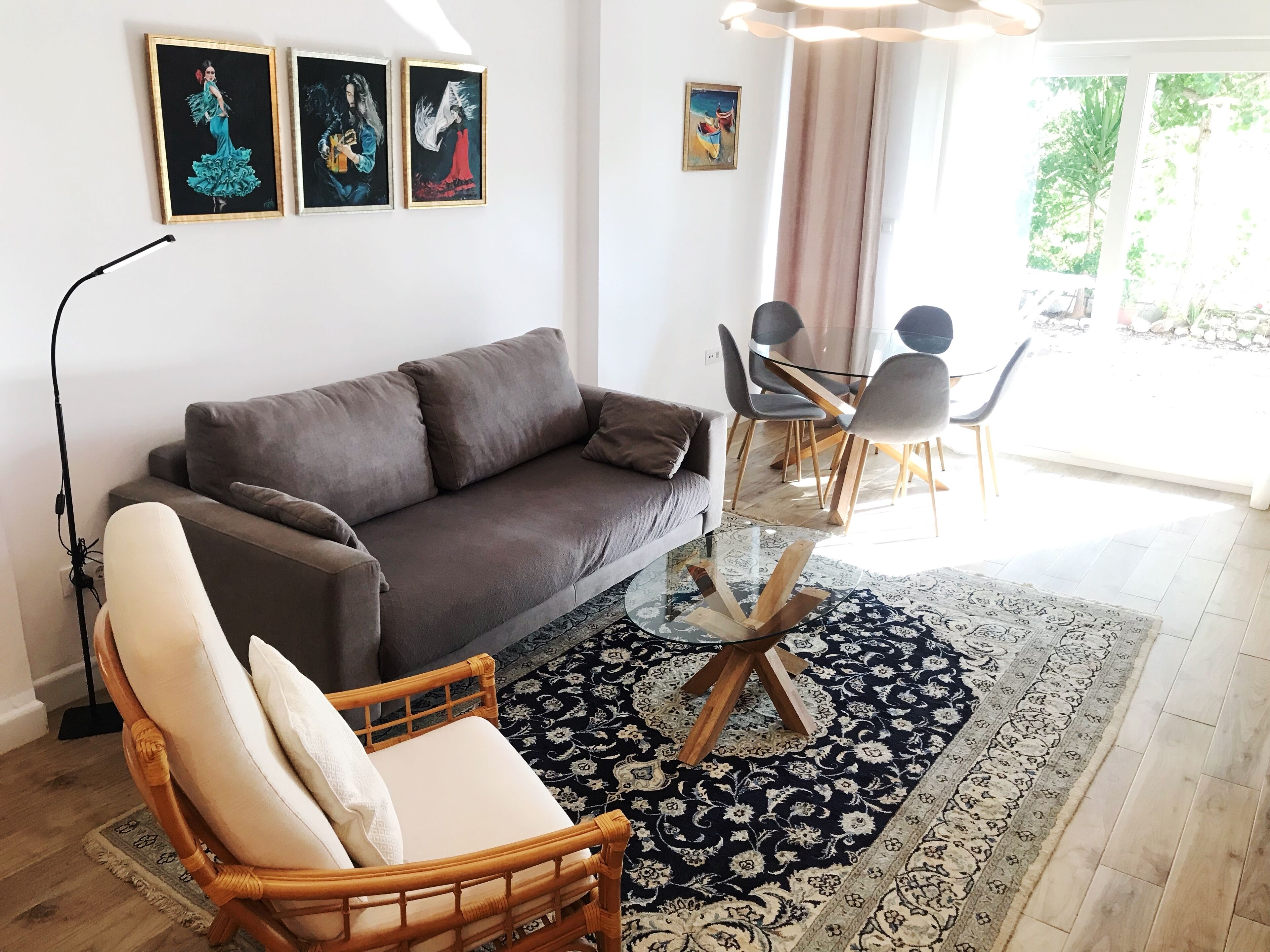 Ein Wohnzimmer mit grauem Sofa, einem Holzstuhl, einem Glastisch und einem gemusterten Teppich. An der Wand hängen drei gerahmte Kunstwerke.