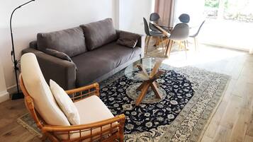 Ein Wohnzimmer mit grauem Sofa, einem Holzstuhl, einem Glastisch und einem gemusterten Teppich. An der Wand hängen drei gerahmte Kunstwerke.