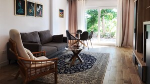 Un salon moderne avec un canapé gris, une chaise en bois, une table basse en verre et un tapis à motifs. Des œuvres d’art encadrées ornent le mur, et une grande fenêtre est agrémentée de rideaux.