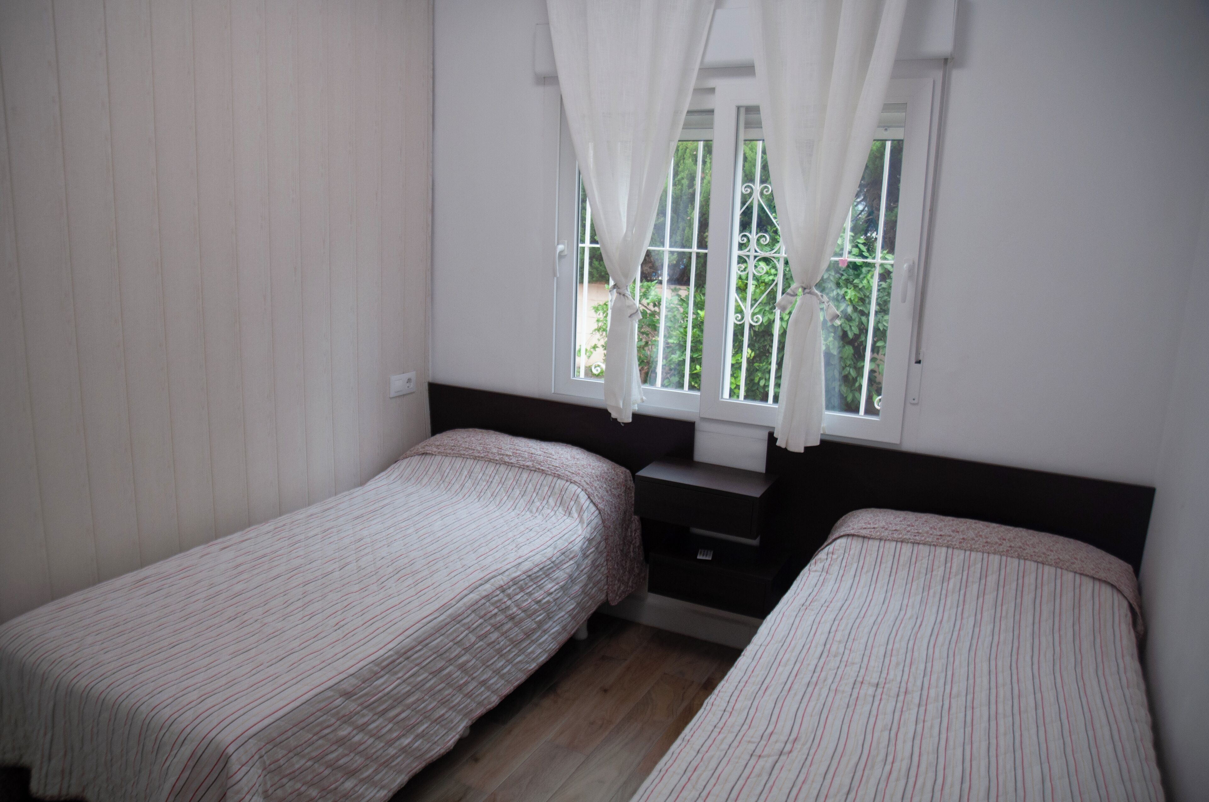 A bedroom with two single beds, a window with curtains, a small bedside table, and a wooden floor.