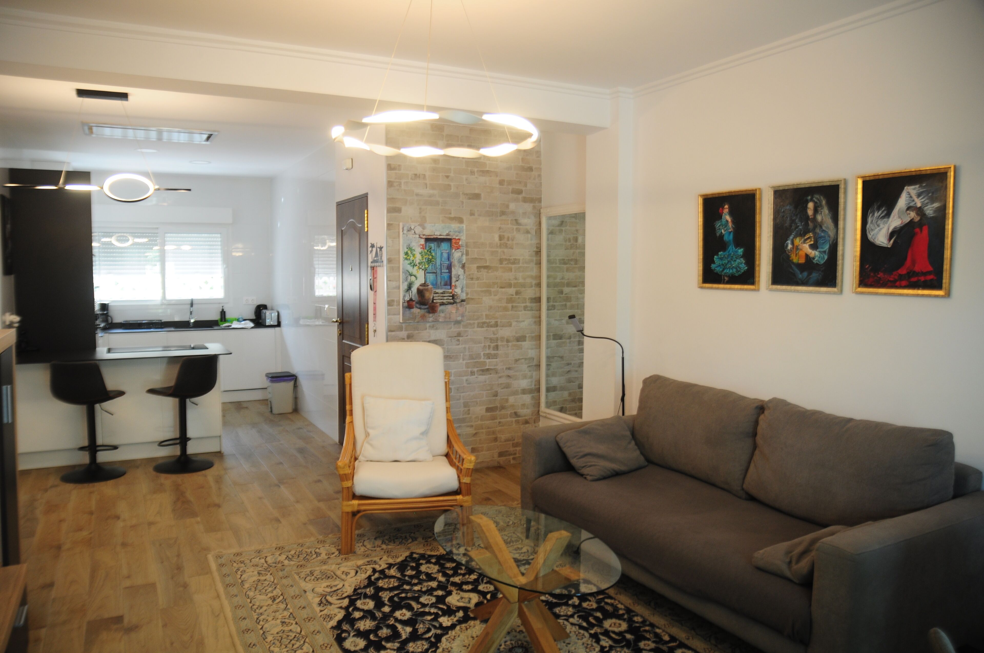 Una sala de estar moderna con un sofá gris, una silla de madera y una mesa de centro de vidrio. Se observa una zona de cocina con encimera blanca y taburetes de bar. En el fondo, una pared de ladrillo y un cuadro.