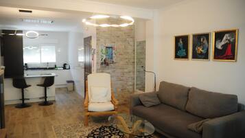 Una sala de estar moderna con un sofá gris, una silla de madera y una mesa de centro de vidrio. Se observa una zona de cocina con encimera blanca y taburetes de bar. En el fondo, una pared de ladrillo y un cuadro.