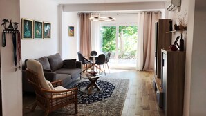 Een moderne woonkamer met een grijze bank, een houten stoel, een salontafel en een eethoek met stoelen en een glazen tafel. Er is een groot raam met gordijnen, een houten kast en kunstwerken aan de muur.