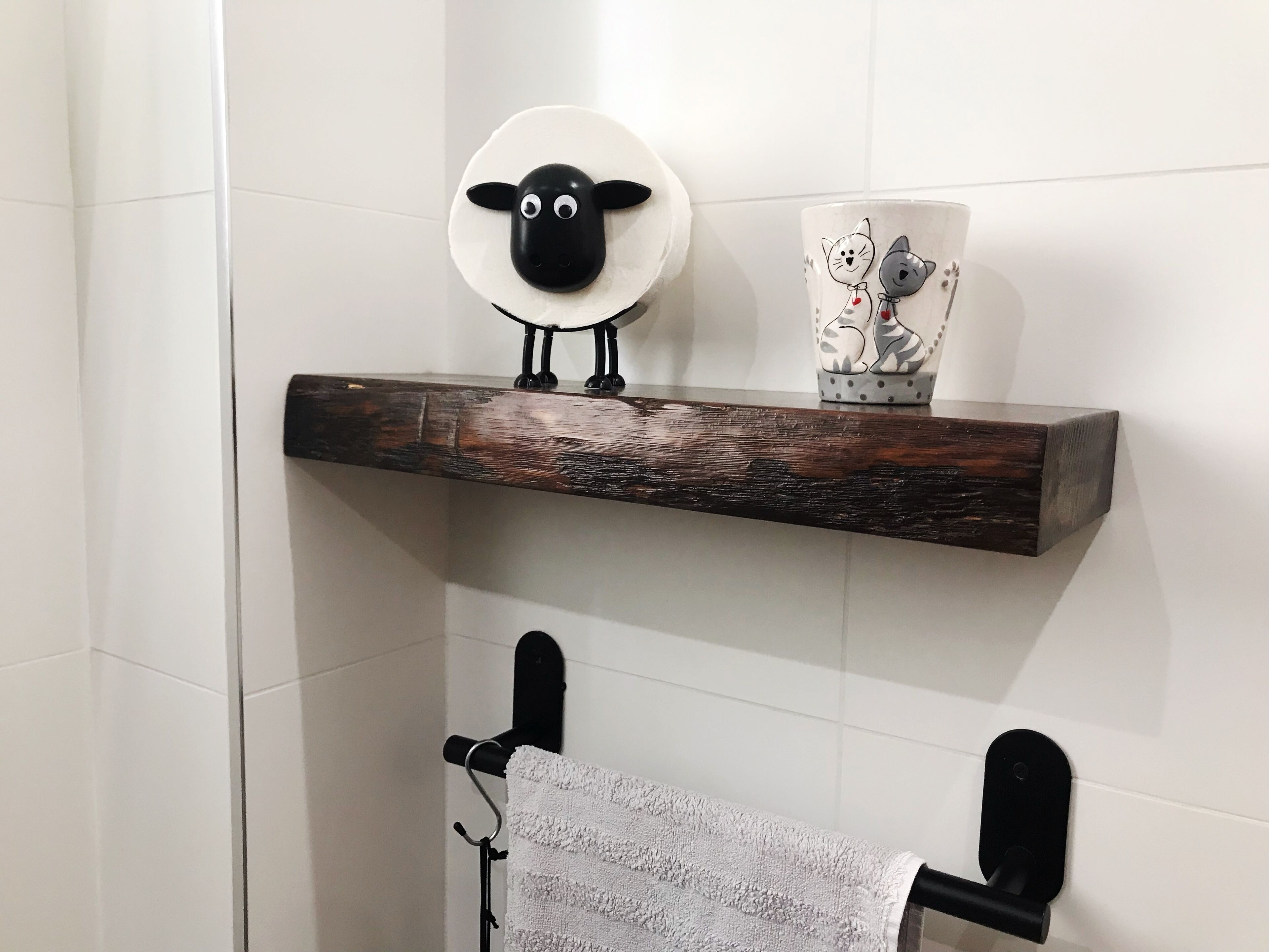 A bathroom with a wooden shelf holding a ceramic sheep head and a mug with cat designs.