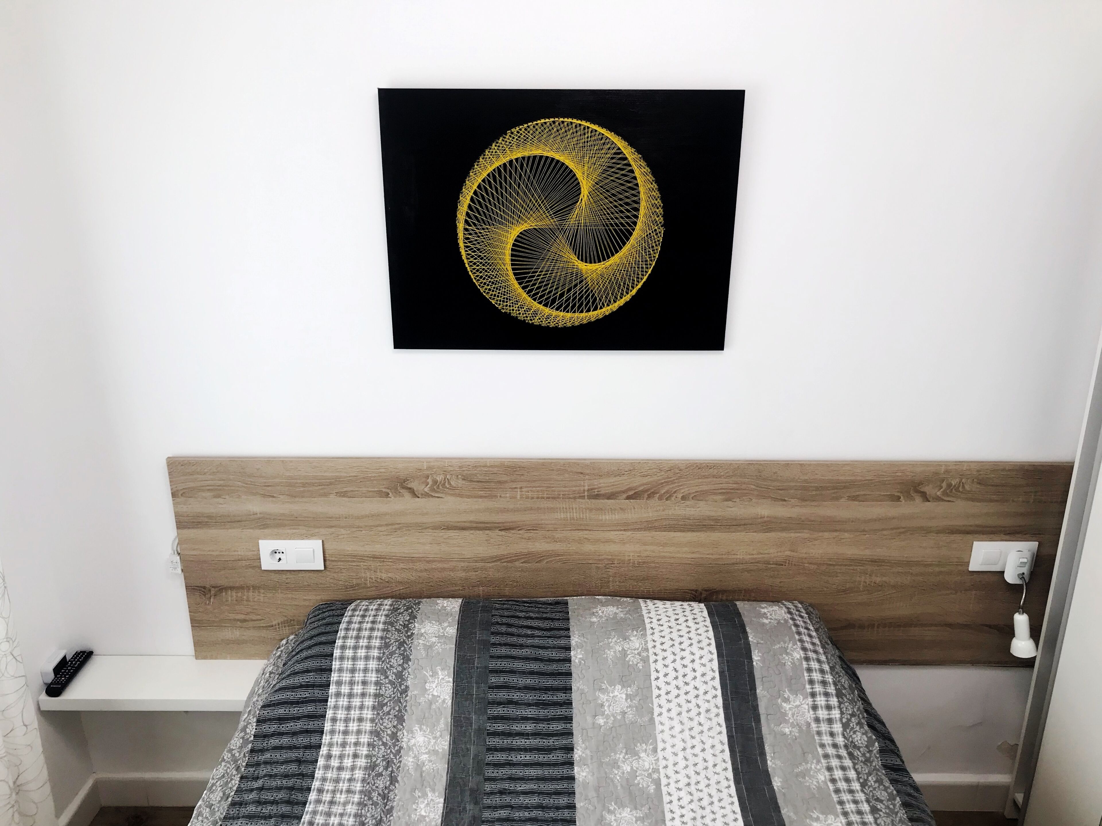 Ein Schlafzimmer mit einem hölzernen Kopfende, einem Bett mit einem gemusterten Bettbezug, einem an der Wand hängenden Kunstwerk und einem Nachttisch mit einer Fernbedienung.