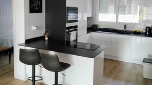 Een moderne keuken met een centraal eiland, twee barkrukken, een inbouwoven en een raam met zonwering.