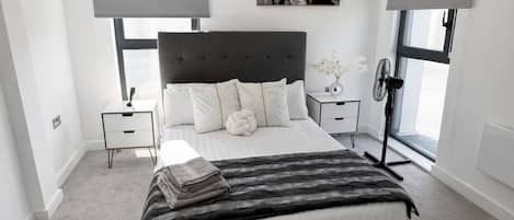 A modern bedroom with a large bed, a bedside table, a nightstand, a floor lamp, and a window with blinds.