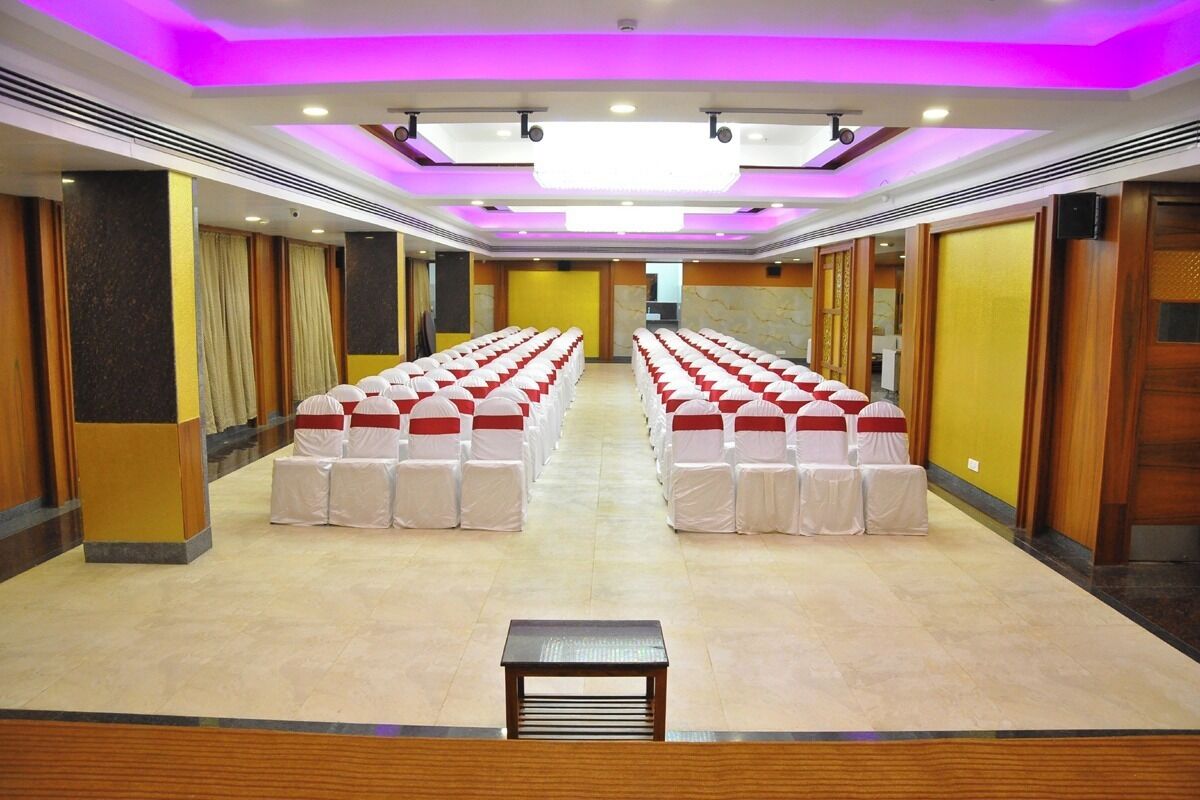 A conference room with rows of chairs covered in white and red sashes, a wooden table in the foreground, and a ceiling with purple lighting.