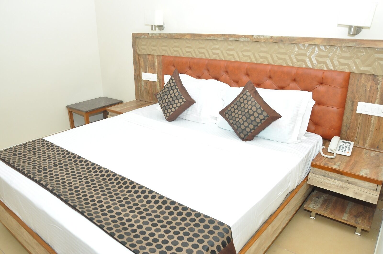 Una cama bien hecha con una colcha estampada, dos almohadas decorativas, una mesita de noche de madera con un teléfono, y una pequeña mesa al lado de la cama.