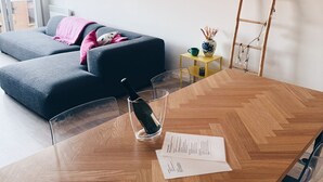 Ein modernes Wohnzimmer mit einem Holzesstisch, einem Glastisch und einer Couch. An der Wand befinden sich gerahmte Bilder und eine Leiter mit Lichterketten.