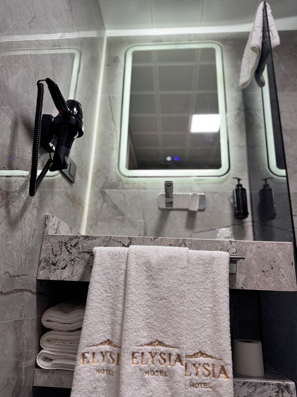 A bathroom with a marble countertop, a towel rack with folded towels, a handheld showerhead, and a window with a view of the outside. - Elysia Hotel (Istanbul)