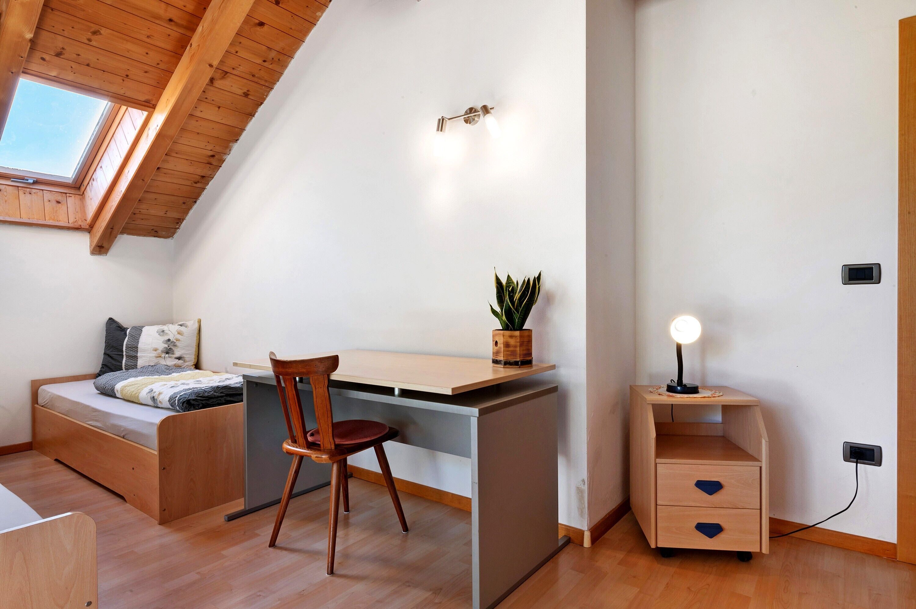 Een compacte kamer met een schuine kap, een bed, een bureau, een stoel, een klein tafeltje met een plant, een nachtkastje met een lamp en een stopcontact.