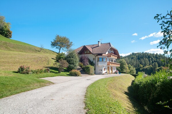 A two-story house with a gravel driveway, surrounded by greenery and hills.
