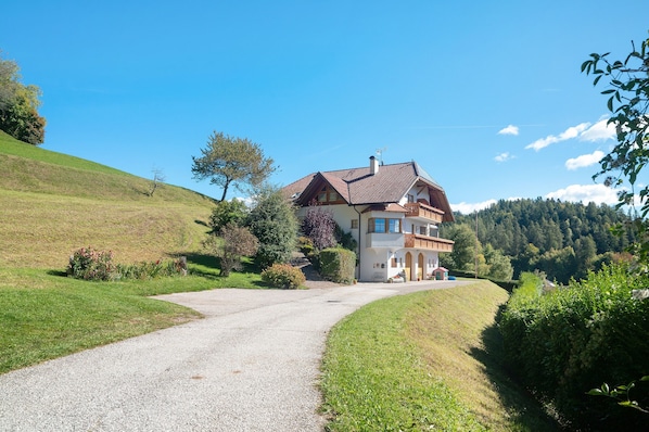 Une maison à deux étages avec une allée en gravier, entourée de verdure et de collines.