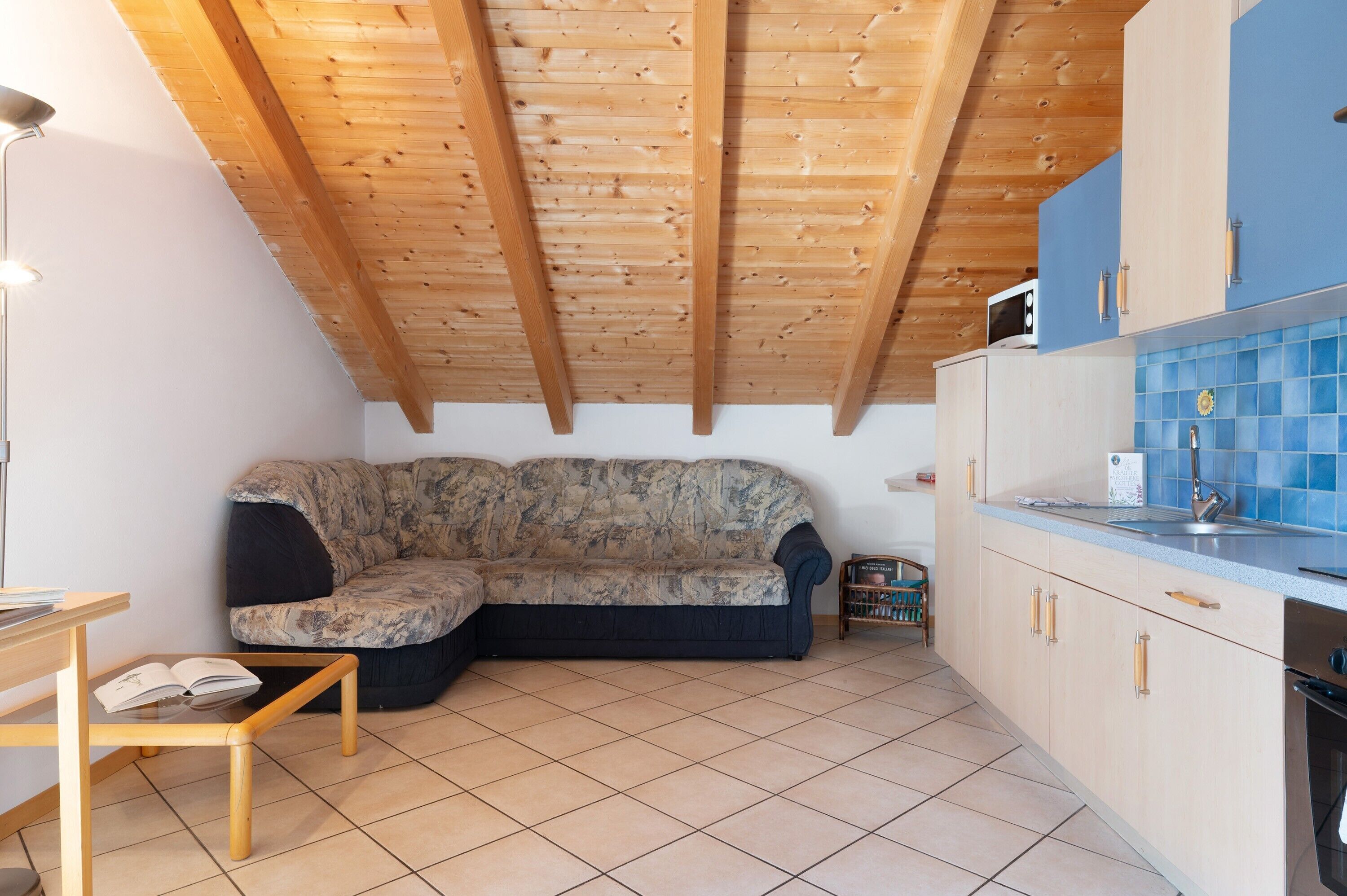 Habitación con techo de madera, un sofá, una zona de cocina con armarios y un microondas, y suelo de baldosas.
