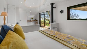 Ein modernes Schlafzimmer mit einem großen Bett, Blick ins Freie und einer kleinen Küchenzeile im Hintergrund.