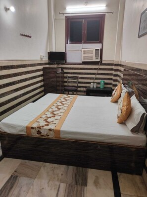 A bedroom with a bed, a chair, a desk, a television, and a window with an air conditioner.