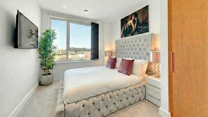 A bedroom with a large bed, a window, a potted plant, and a painting on the wall.