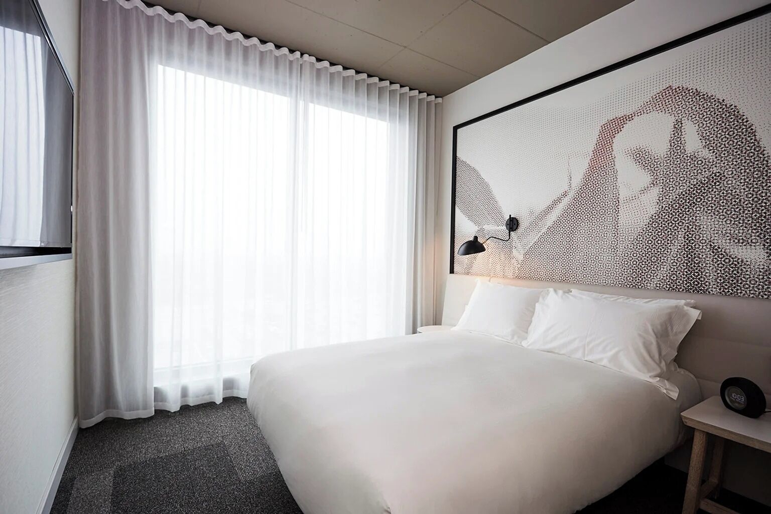 Une chambre d’hôtel avec un grand lit, une fenêtre avec des rideaux légers, une œuvre d’art accrochée au mur et une table de chevet avec une lampe.