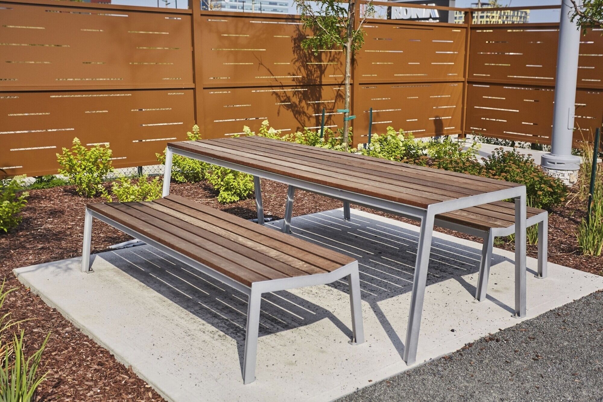 Two wooden picnic tables with metal frames, set on a concrete surface with mulch and small plants around.